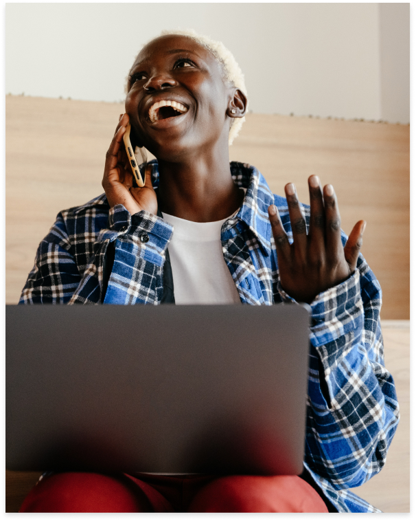 A imagem mostra uma mulher negra de camisa xadrez azul sorrindo falando no celular e com o notebook no colo.